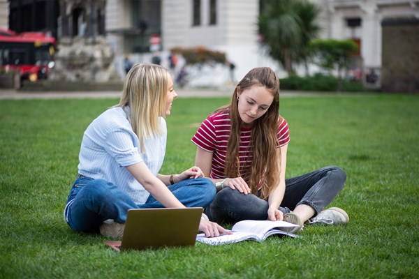 Two female students sitting in the park studying together with a book and a laptop.
