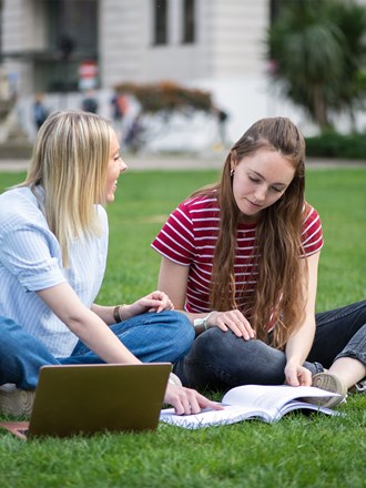 Two female students sitting in the park studying together with a book and a laptop.